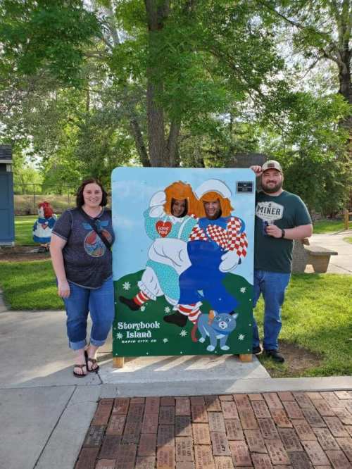 Three people pose behind a colorful cutout at Storybook Island, smiling and holding drinks in a sunny park setting.