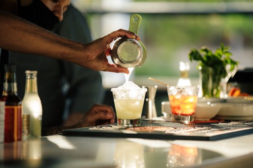 A bartender pours a cocktail into a glass filled with ice, with fresh ingredients and another drink in the background.