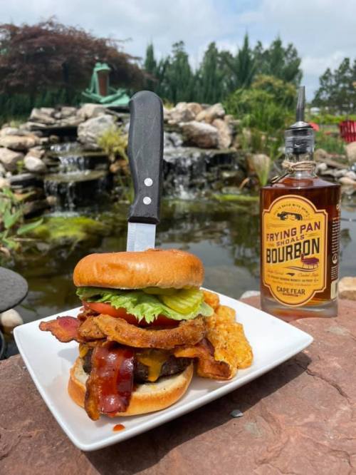 A gourmet burger topped with bacon, lettuce, and tomato, served on a plate beside a bottle of bourbon and a scenic pond.