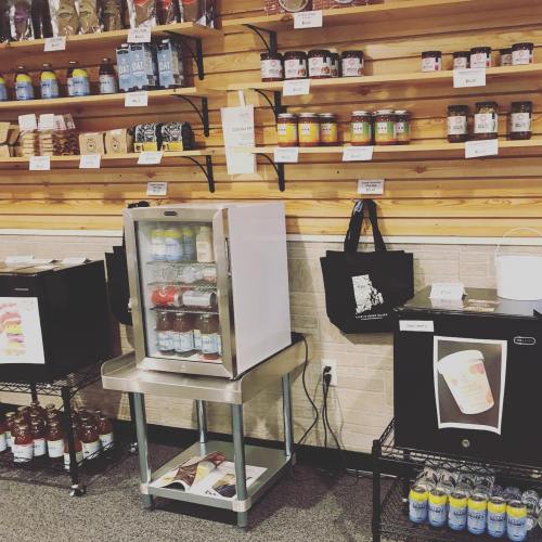 A display of shelves filled with jars and drinks, featuring a small fridge and a black cooler on a metal stand.