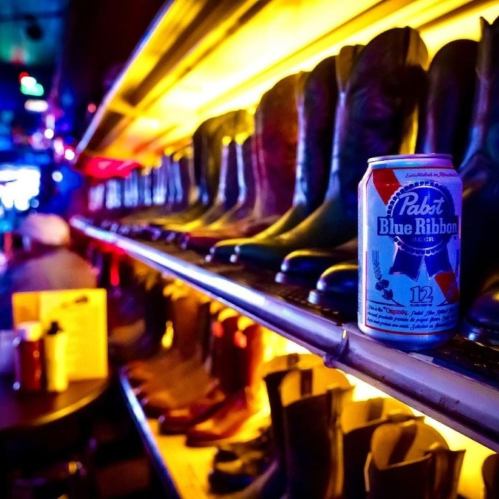 A row of cowboy boots on a shelf with a can of Pabst Blue Ribbon beer in the foreground, illuminated by colorful lights.