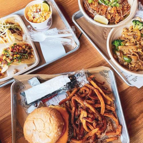 A wooden table with trays of food: tacos, noodles, a burger, and sweet potato fries.