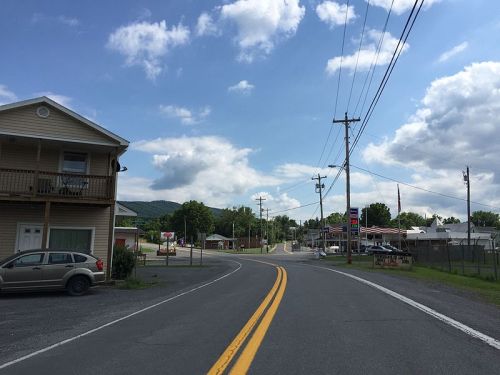 A rural road curves past a small town with buildings, power lines, and a blue sky dotted with clouds.