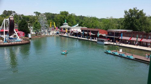 A scenic view of a park with a lake, amusement rides, and people enjoying the attractions on a sunny day.