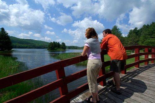 A woman and man stand on a red bridge overlooking a serene lake surrounded by trees and clouds.