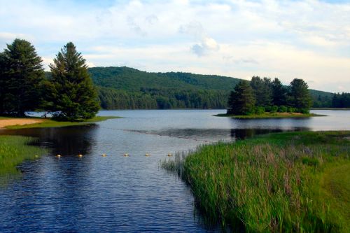 A serene lake surrounded by trees and hills, with calm waters and grassy banks under a partly cloudy sky.