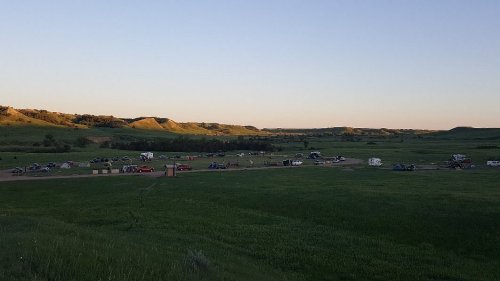 A wide view of a grassy campsite with various tents and RVs, surrounded by rolling hills at sunset.