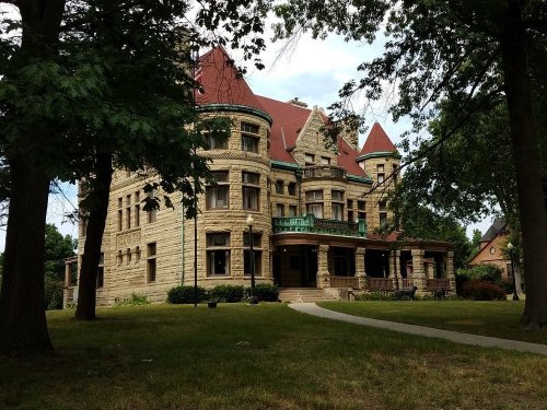 A large, historic stone mansion with a red roof, surrounded by trees and a grassy lawn.