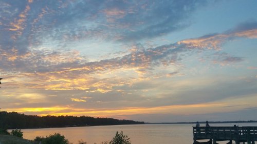A serene sunset over a calm lake, with colorful clouds reflecting on the water and a wooden pier in the foreground.