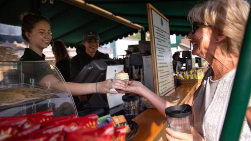A woman receives a cupcake from a barista at a café, with a young man in the background and snacks on display.