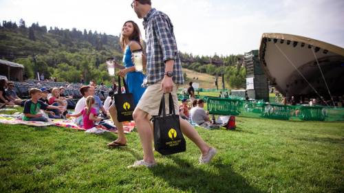 A couple walks on grass carrying bags, enjoying a sunny outdoor event with a stage and audience in the background.