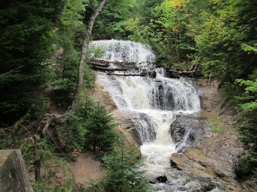 A serene waterfall cascades down rocky steps, surrounded by lush green trees and vibrant autumn foliage.