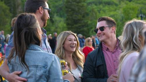 A group of friends laughing and chatting outdoors at a lively gathering, surrounded by greenery.