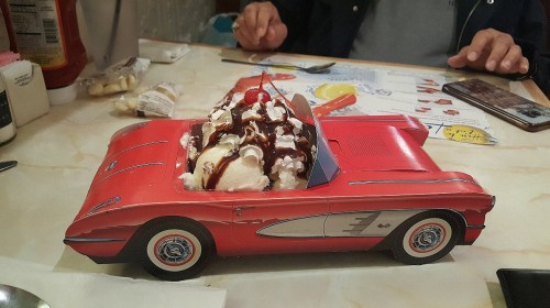 A red toy car filled with ice cream, whipped cream, and toppings, placed on a table with utensils nearby.