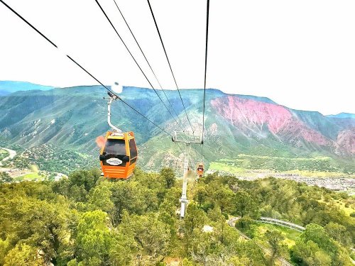 A scenic view of a cable car traveling over green mountains with a valley below and colorful rock formations in the distance.
