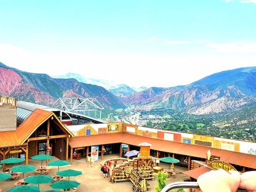 A scenic view from a mountain overlook, featuring a valley, river, and colorful hills with a rustic marketplace below.