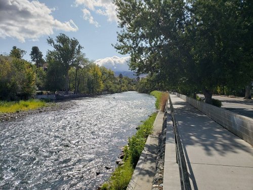 A serene river flows beside a paved path, lined with trees and greenery under a partly cloudy sky.