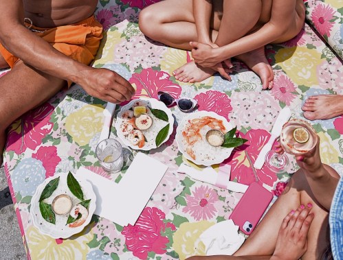 A colorful picnic scene with people enjoying seafood dishes and drinks on a floral-patterned blanket.