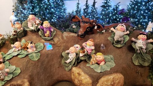 A whimsical display featuring dolls on large leaves, surrounded by a cow and decorated with twinkling lights and trees.