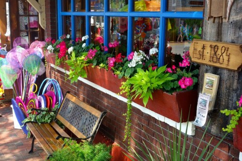 Colorful flower boxes with pink and white blooms adorn a brick storefront, alongside a bench and playful outdoor items.