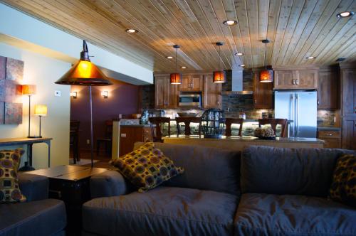 Cozy living room with a sofa, wooden ceiling, and a modern kitchen in the background featuring warm lighting and wooden cabinets.
