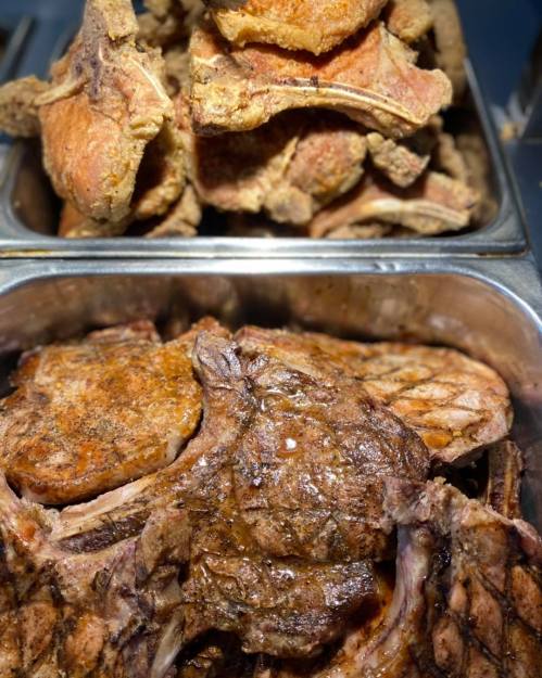 A close-up of cooked meat chops in a metal tray, with a stack of additional chops in the background.