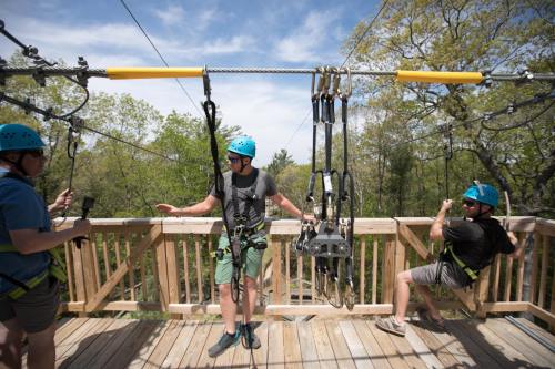 Three men in helmets prepare for a zipline adventure on a wooden platform surrounded by trees.