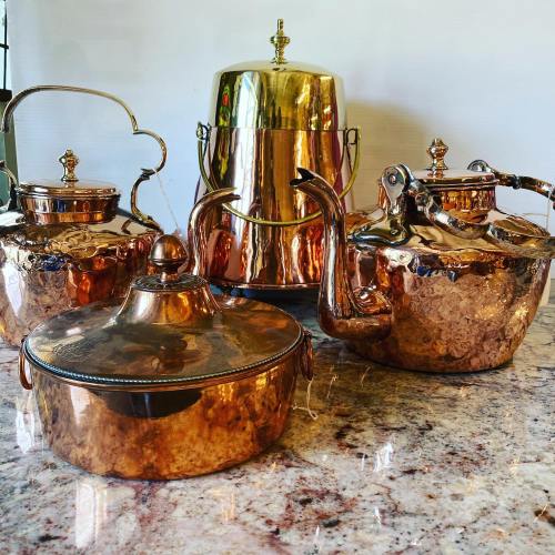 A collection of polished copper kitchenware, including kettles and a pot, displayed on a marble surface.