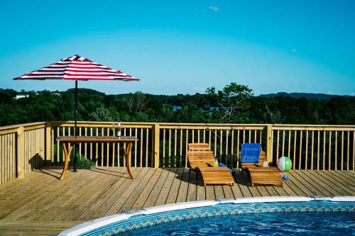 A sunny poolside deck with a striped umbrella, wooden chairs, and a table, surrounded by greenery and blue sky.