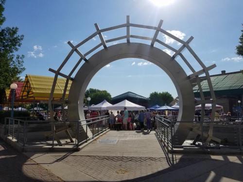 A large metal arch frames a bustling market scene with tents and people under a clear blue sky.