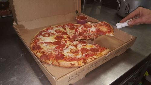 A hand pulls a slice of pepperoni pizza from an open pizza box, with cheese stretching and a small cup of sauce on the side.