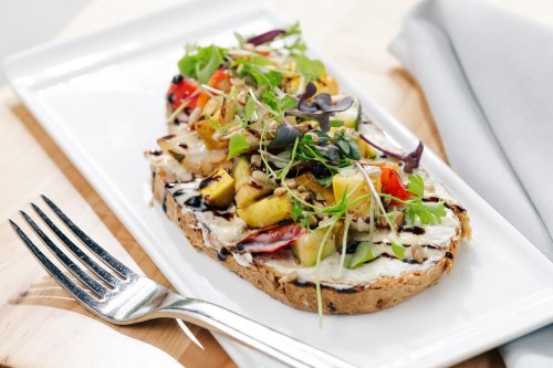 A slice of toasted bread topped with grilled vegetables and microgreens, served on a white plate with a fork.