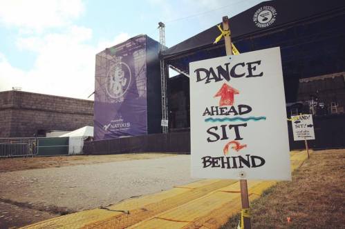 Sign at a festival directing attendees: "Dance Ahead" with an arrow up and "Sit Behind" with an arrow down. Stage in background.