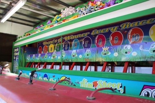 Colorful carnival game booth with numbered racing frogs and plush toys displayed above. Brightly painted and inviting.