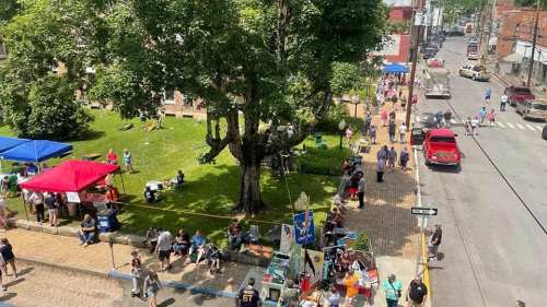 A bustling street fair with tents, vendors, and people enjoying the day under a large tree.
