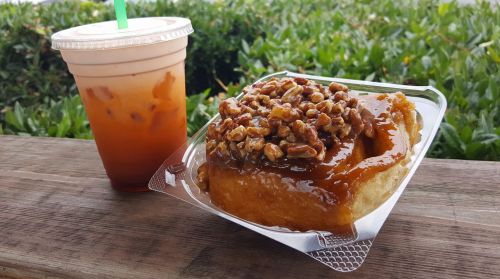A sticky bun topped with nuts next to a cup of iced tea on a wooden surface, with greenery in the background.
