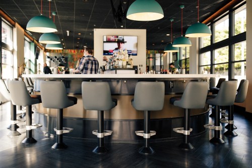 A modern bar with a circular counter, high stools, and a TV in the background, featuring colorful pendant lights.