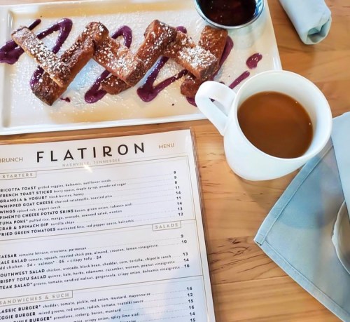 A plate of French toast drizzled with berry sauce, powdered sugar, a cup of coffee, and a menu on a wooden table.