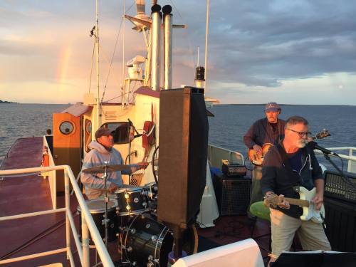 Three musicians perform on a boat, with a rainbow in the background and a scenic water view.