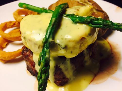 A plated steak topped with hollandaise sauce and asparagus, served with a baked potato and onion rings.