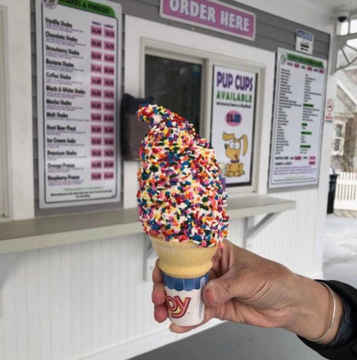 A hand holds a colorful ice cream cone topped with sprinkles, in front of an ice cream stand menu.