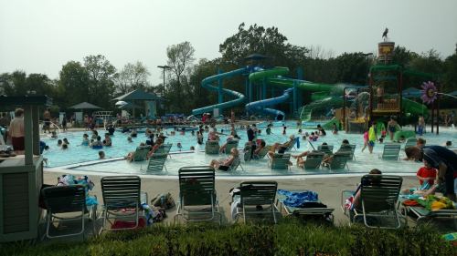 A busy outdoor pool scene with people swimming, lounging on chairs, and water slides in the background.