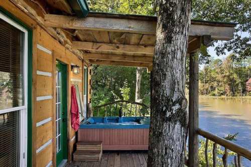 A cozy cabin porch with a hot tub overlooking a serene lake, surrounded by trees and nature.