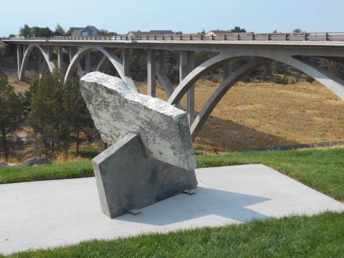 A large abstract stone sculpture on a concrete base, with a bridge and grassy landscape in the background.