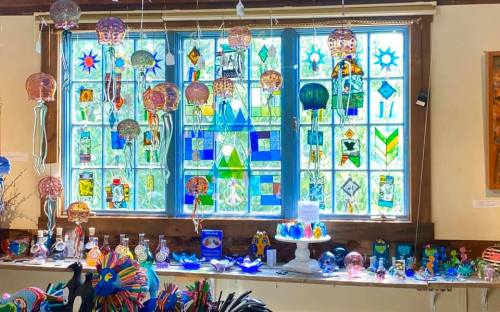 Colorful stained glass windows with various glass art pieces displayed on a table below, creating a vibrant atmosphere.