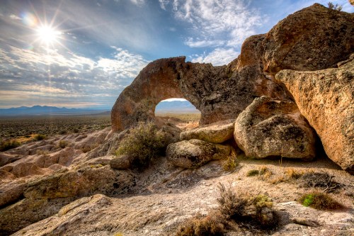 A rocky landscape features a natural arch under a bright sky with scattered clouds and distant mountains.