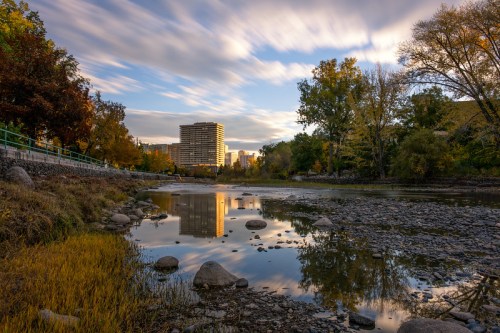 A serene river scene with reflections of buildings and trees, surrounded by autumn foliage under a cloudy sky.