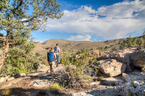 Two hikers walking on a rocky trail surrounded by trees and mountains under a partly cloudy sky.