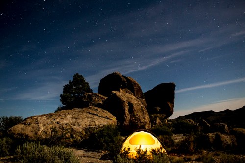 A glowing tent sits among large rocks under a starry night sky, surrounded by sparse vegetation.
