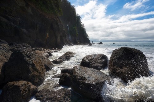 Rocky coastline with crashing waves, lush cliffs, and a cloudy blue sky in the background.